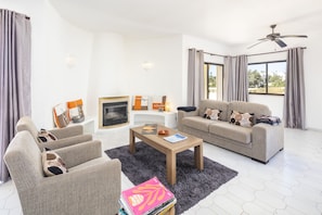 A living room with a sofa, armchair, coffee table, fireplace, and large windows with curtains. - Casa San Michel - holiday villa with private pool, air-conditioning and sea view (Lagoa)