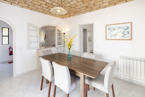 A dining area with a wooden table, six chairs, a vase with flowers, a kitchen in the background, and a framed picture on the wall.