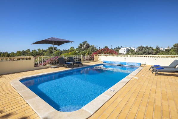 A rectangular swimming pool with a tiled edge, surrounded by a tiled deck. There are lounge chairs and a large umbrella on the deck.