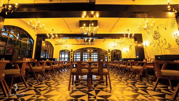 A restaurant with a geometric patterned floor, wooden chairs, chandeliers, and a large arched window.