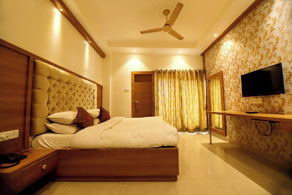 A bedroom with a tufted headboard, a bed with white bedding, a bedside table with a lamp, a ceiling fan, and a TV mounted on the wall.
