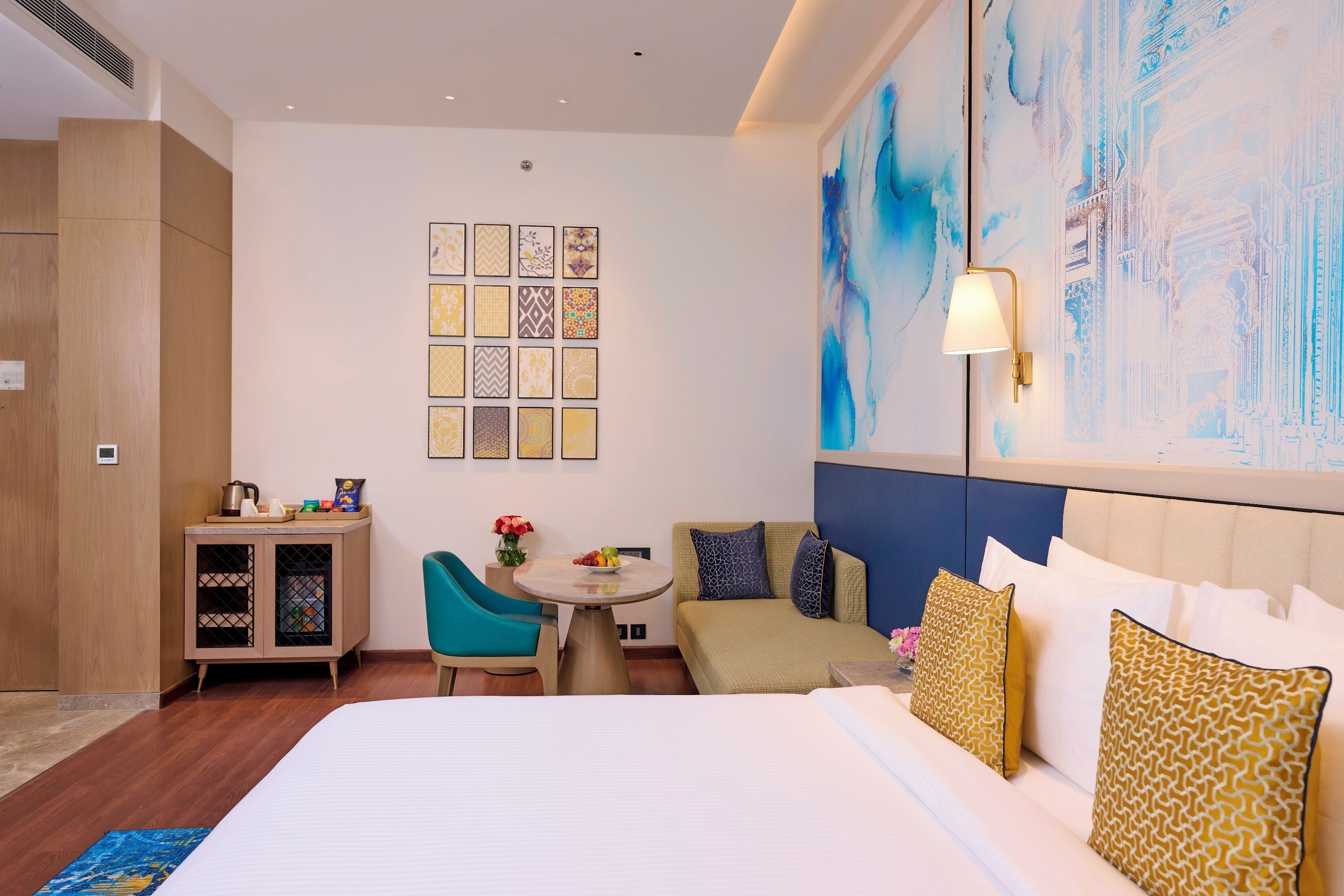 A hotel room with a bed, a desk with a chair, a small table with a vase of flowers, and a wall with a large abstract painting.