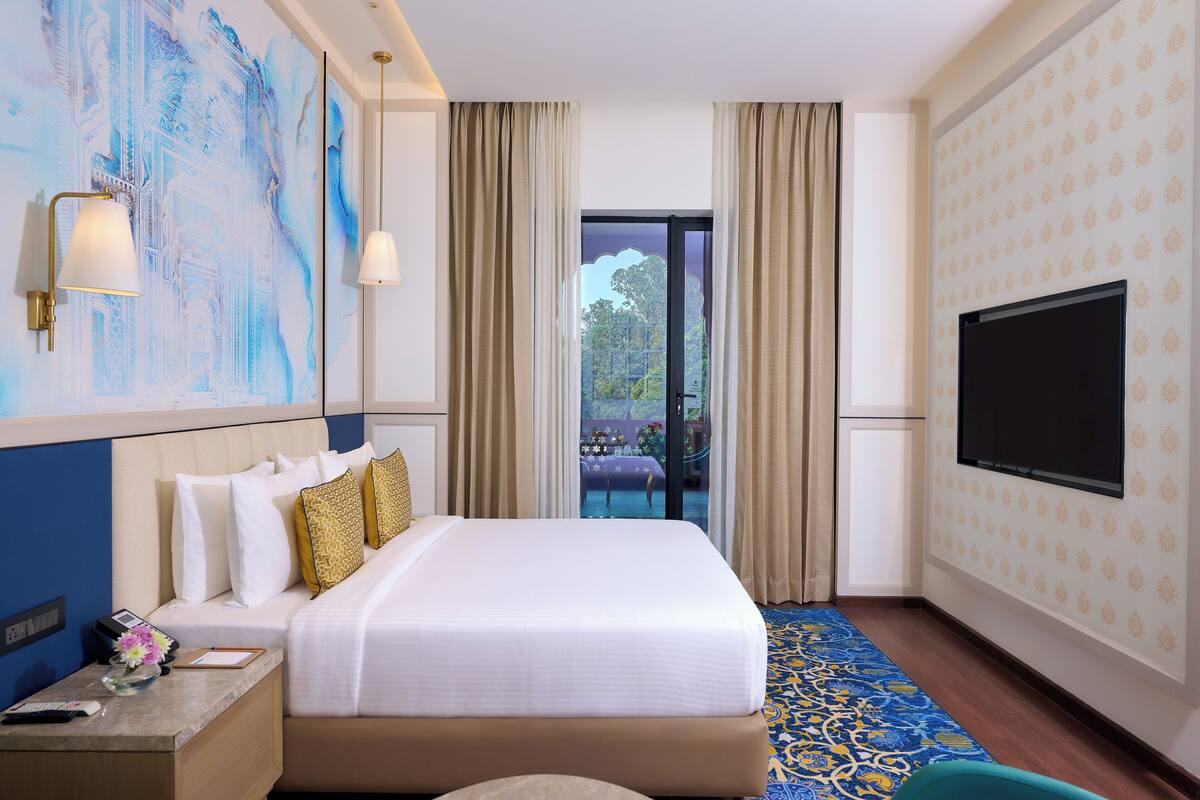 A hotel room with a large bed, a flat-screen TV, a nightstand, a desk, and a view of the outdoors through a glass door.