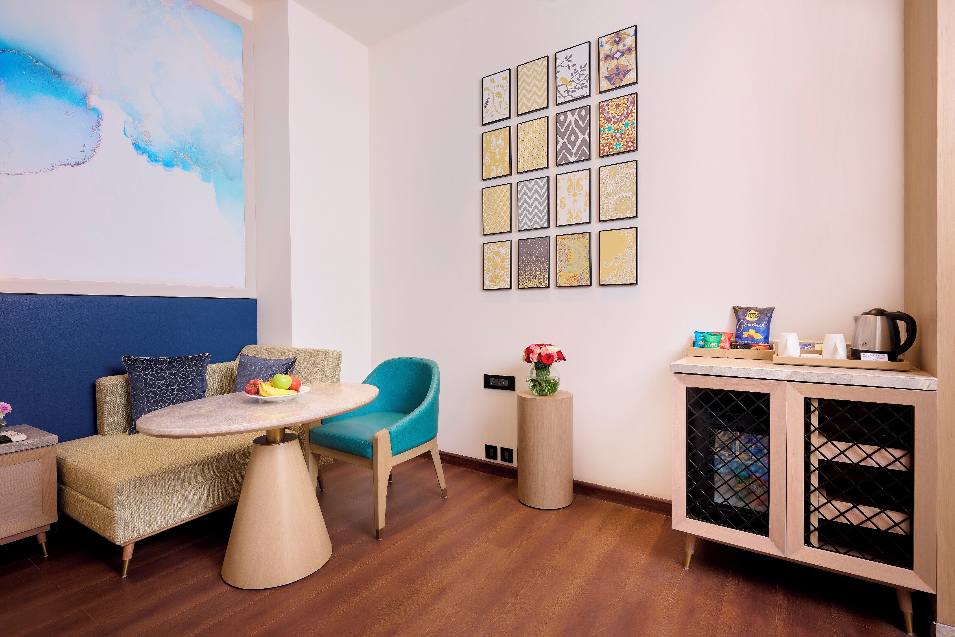 A modern room with a sofa, a blue chair, a round table with fruits, a cabinet with kitchen items, and a wall with framed art.