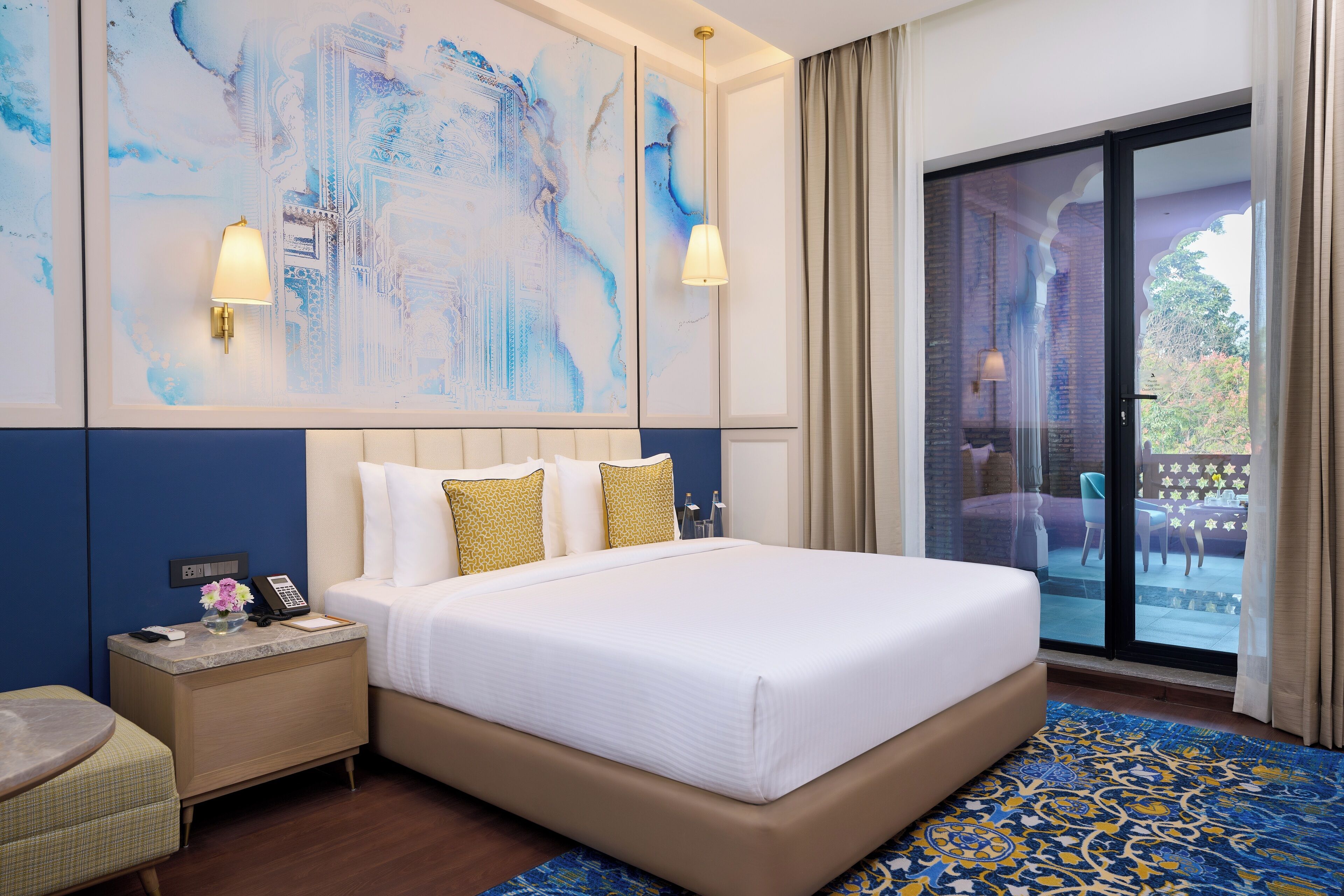 A hotel room with a large bed, a nightstand with a phone and lamp, a view of the outside patio through a sliding glass door, and a large abstract painting on the wall.