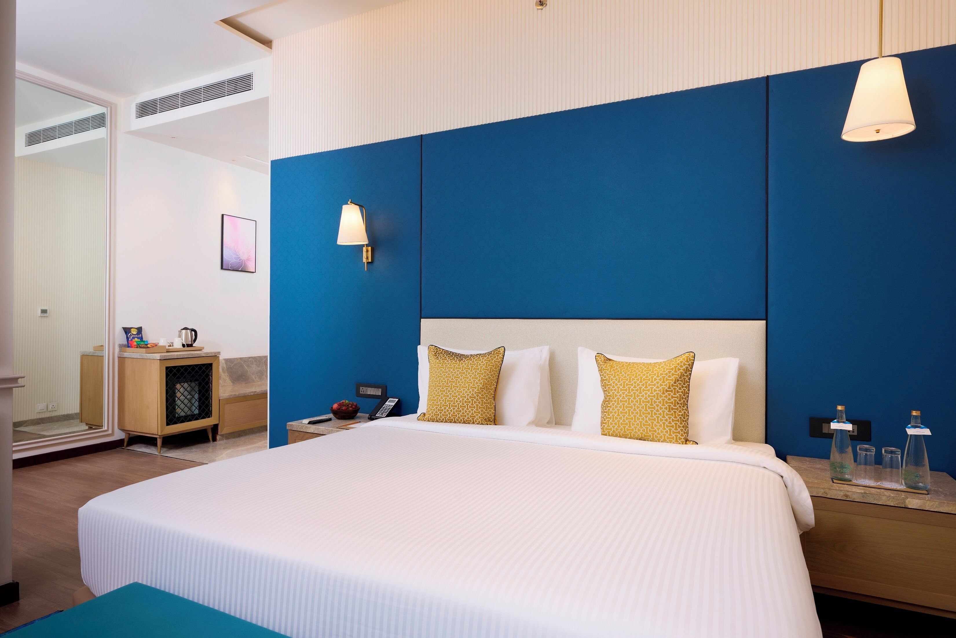 Une chambre d’hôtel avec un grand lit, une table de chevet avec une lampe, une petite armoire en bois et un miroir.