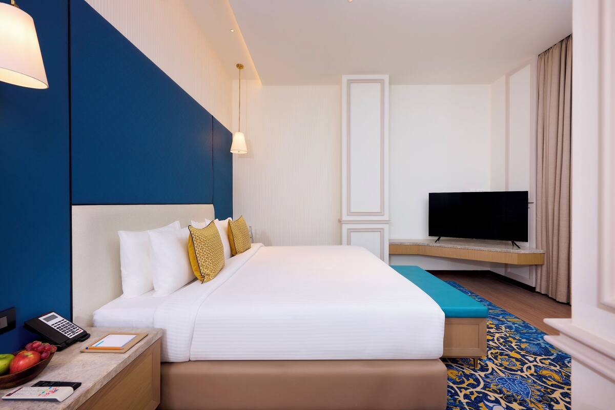 A hotel room with a large bed, a flat-screen TV mounted on the wall, a bedside table with a lamp, and a patterned rug.