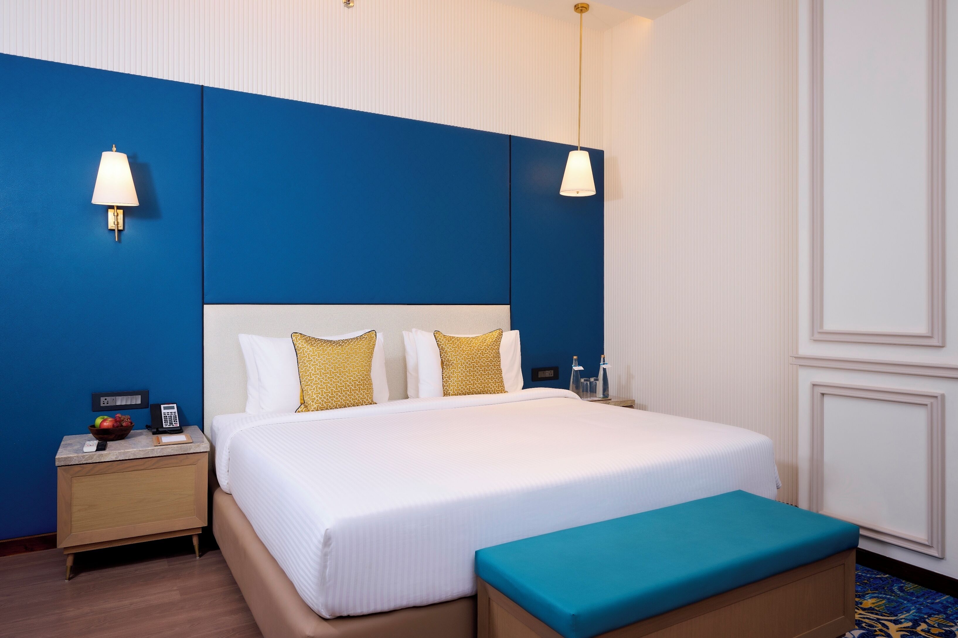 Ein Hotelzimmer mit einem großen Bett, einem Nachttisch mit Lampe, einem Telefon und einer Obstschale.