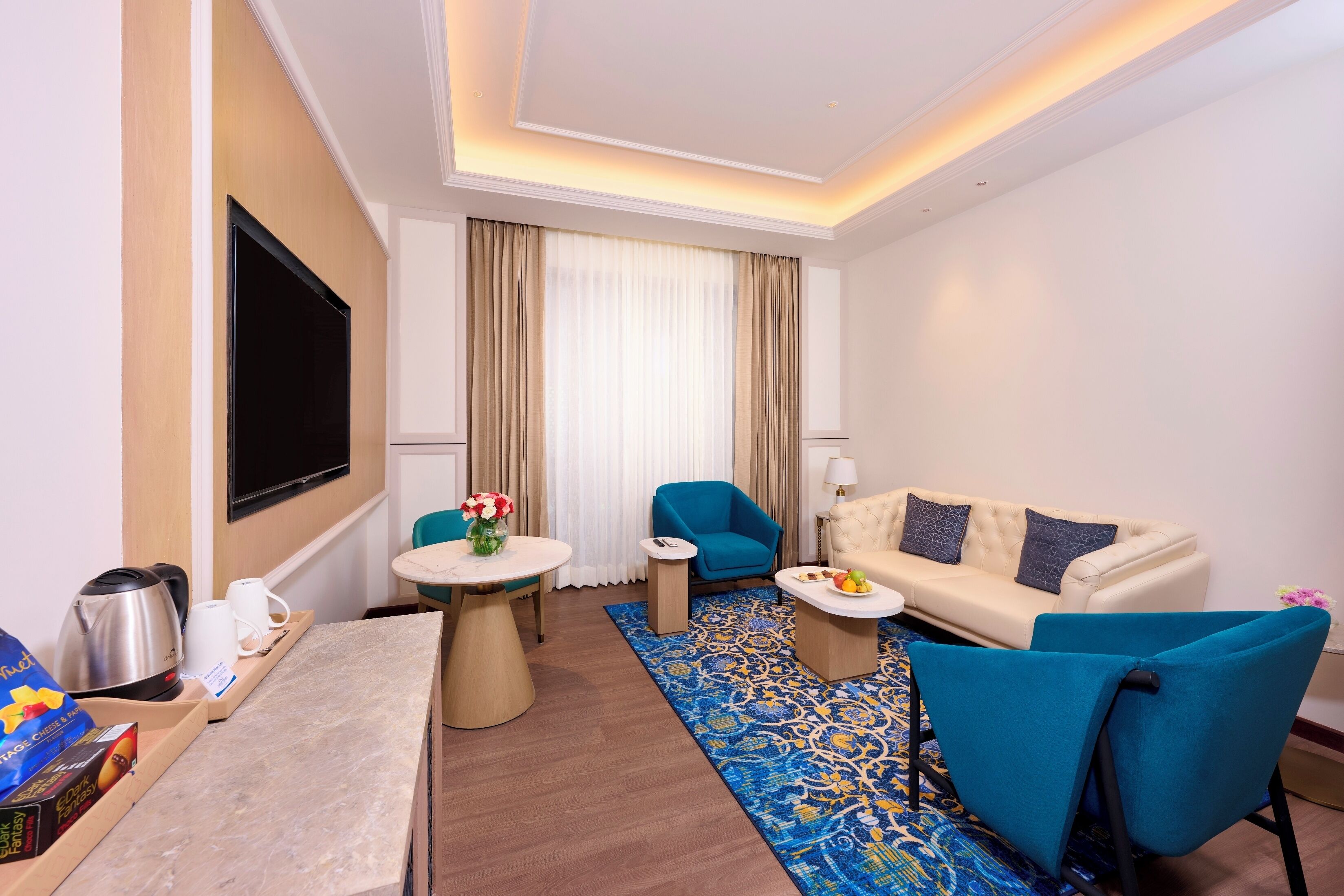 Una habitación de hotel moderna con televisor de pantalla plana, un sofá color beige, un sillón azul, una mesita redonda pequeña y una estación de café.