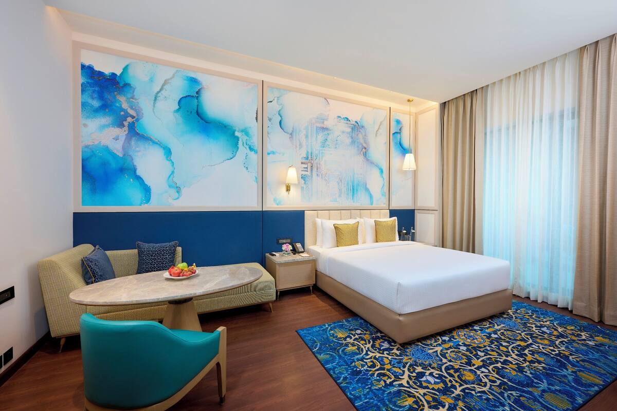 A hotel room with a bed, a sofa, a small table with fruit, and abstract wall art.