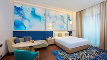 A hotel room with a bed, a sofa, a small table with fruit, and abstract wall art.
