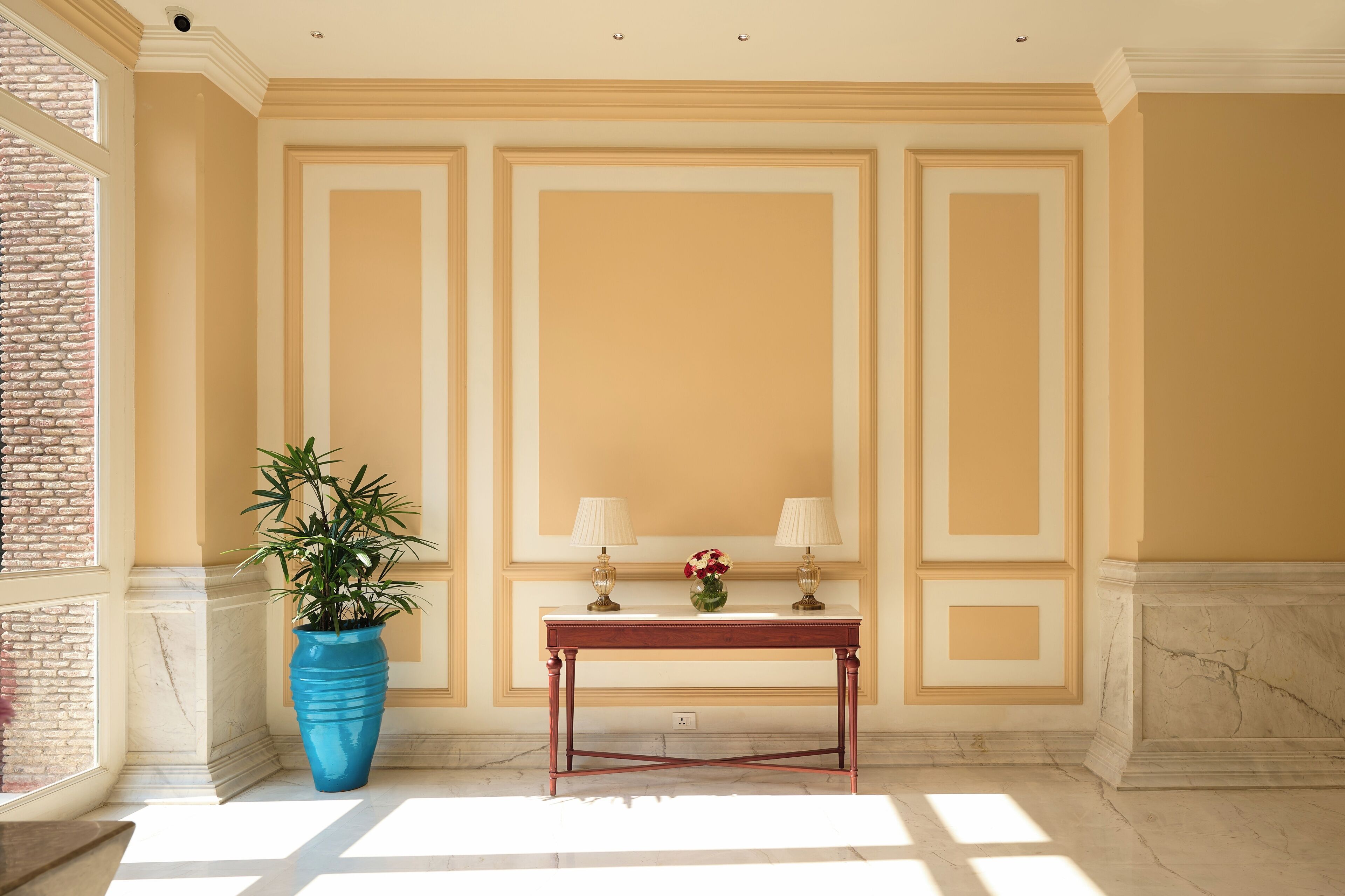 A room with a marble floor, a large blue potted plant, a wooden table with two lamps, and a vase with flowers.