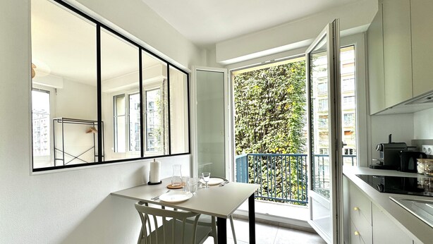 A modern kitchen with a dining area, large windows, and a view of greenery outside.