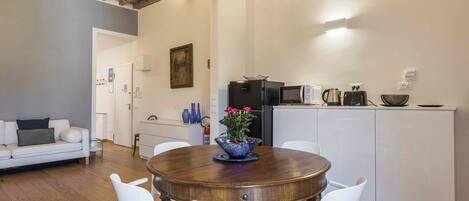 A dining area with a round wooden table, white chairs, a blue vase with flowers, and a kitchen in the background.