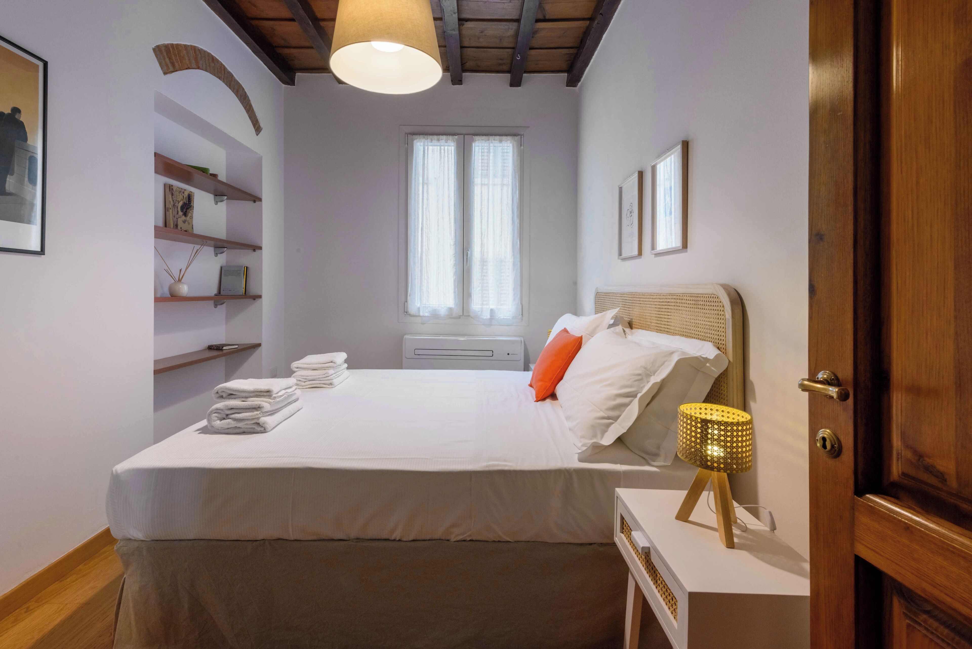 A neatly made bed with white linens, a bedside lamp, and a window with sheer curtains.