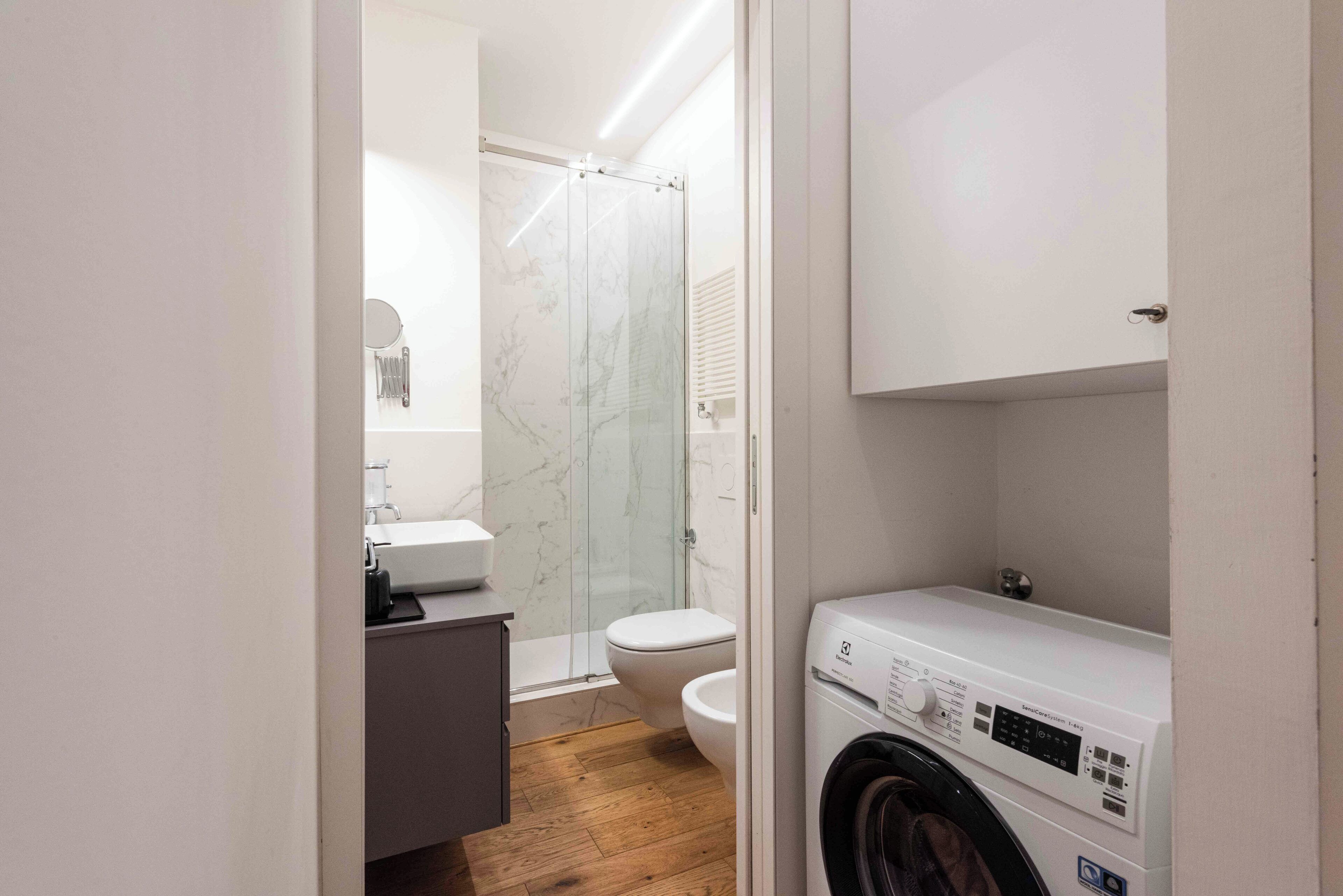 Un baño moderno con cabina de ducha de vidrio, inodoro, lavabo con espejo y lavadora de carga frontal.