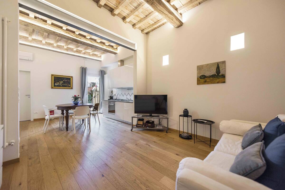 Een ruime woonkamer met houten plafond, een eettafel met stoelen, een bank, een televisie en een keuken op de achtergrond.