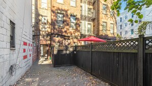 Une ruelle étroite, bordée d’une clôture en bois, avec un parasol rouge et un bâtiment aux murs de briques et aux fenêtres.
