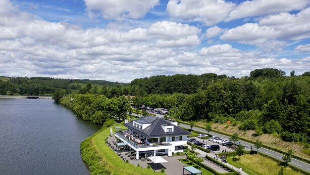 A riverside hotel with outdoor seating, a parking area, and a view of the river and surrounding greenery.