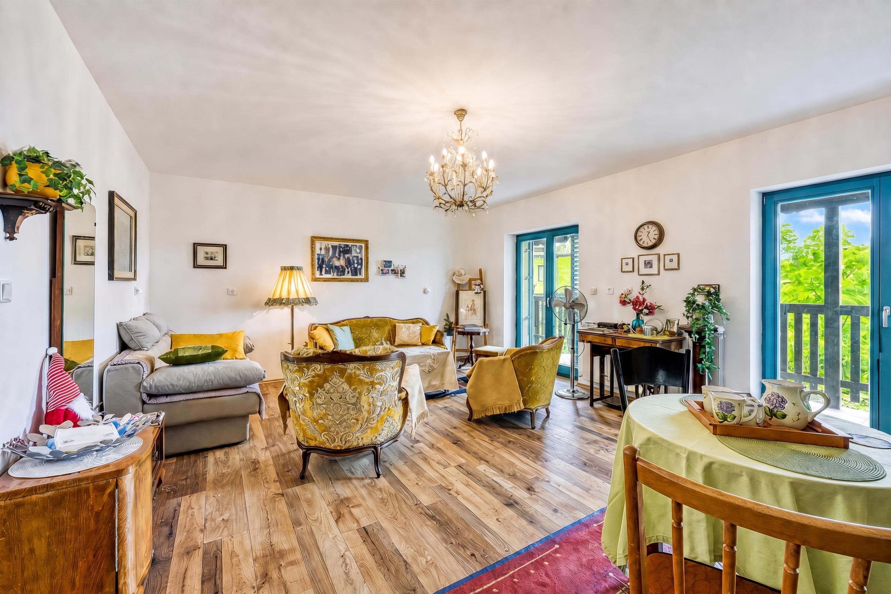 Un'ampia zona giorno con pavimento in legno, un lampadario, un tavolo da pranzo con tovaglia verde e vista attraverso una finestra con cornice blu.