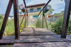 A wooden bridge leading to a house with a balcony, surrounded by greenery.
