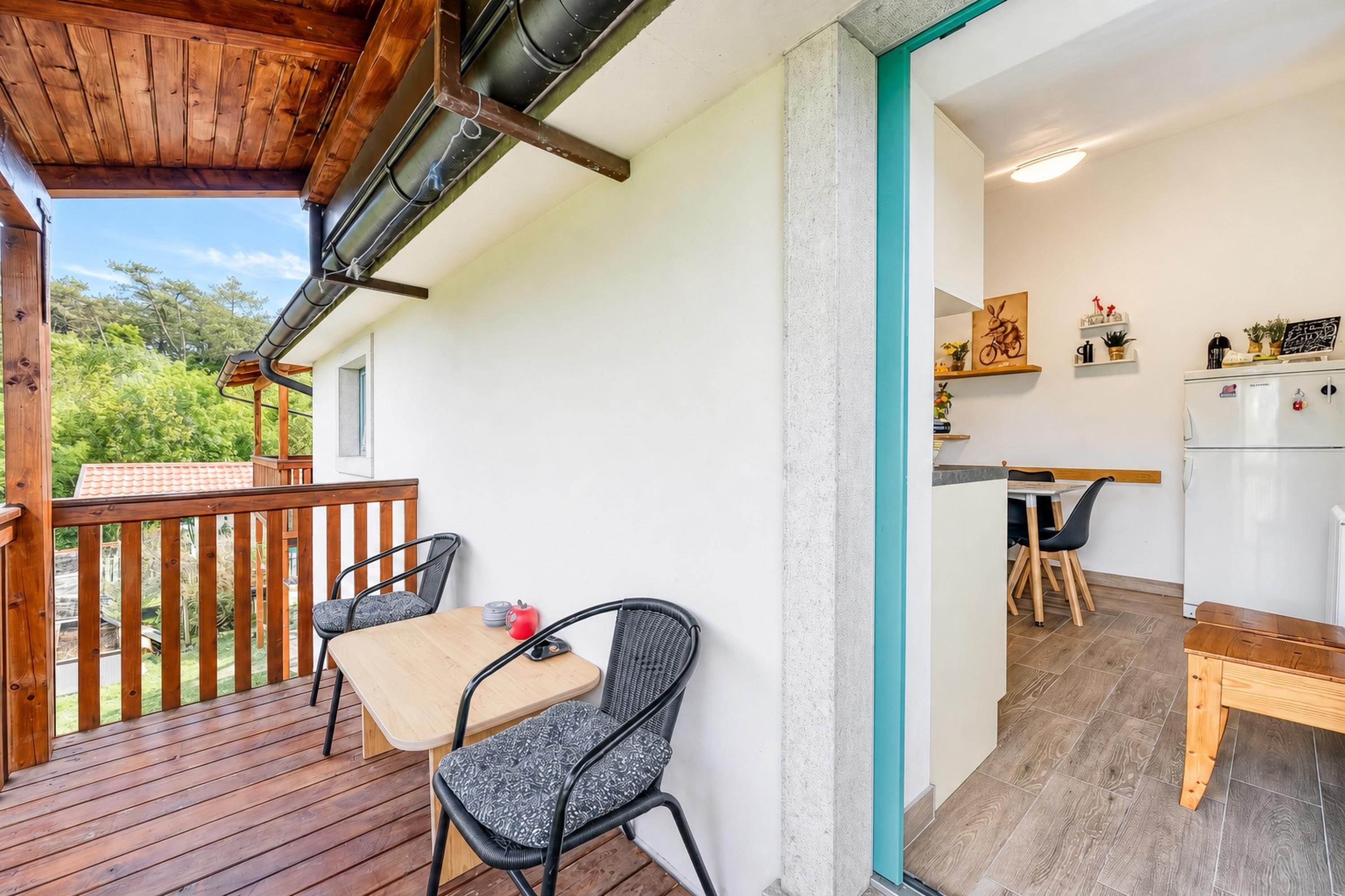 A wooden deck with a table and chairs, a kitchen area with a refrigerator, and a view of greenery.