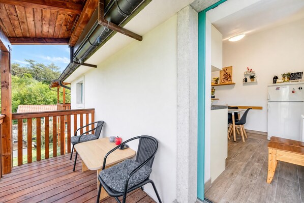 A wooden deck with a table and chairs, a kitchen area with a refrigerator, and a view of greenery.
