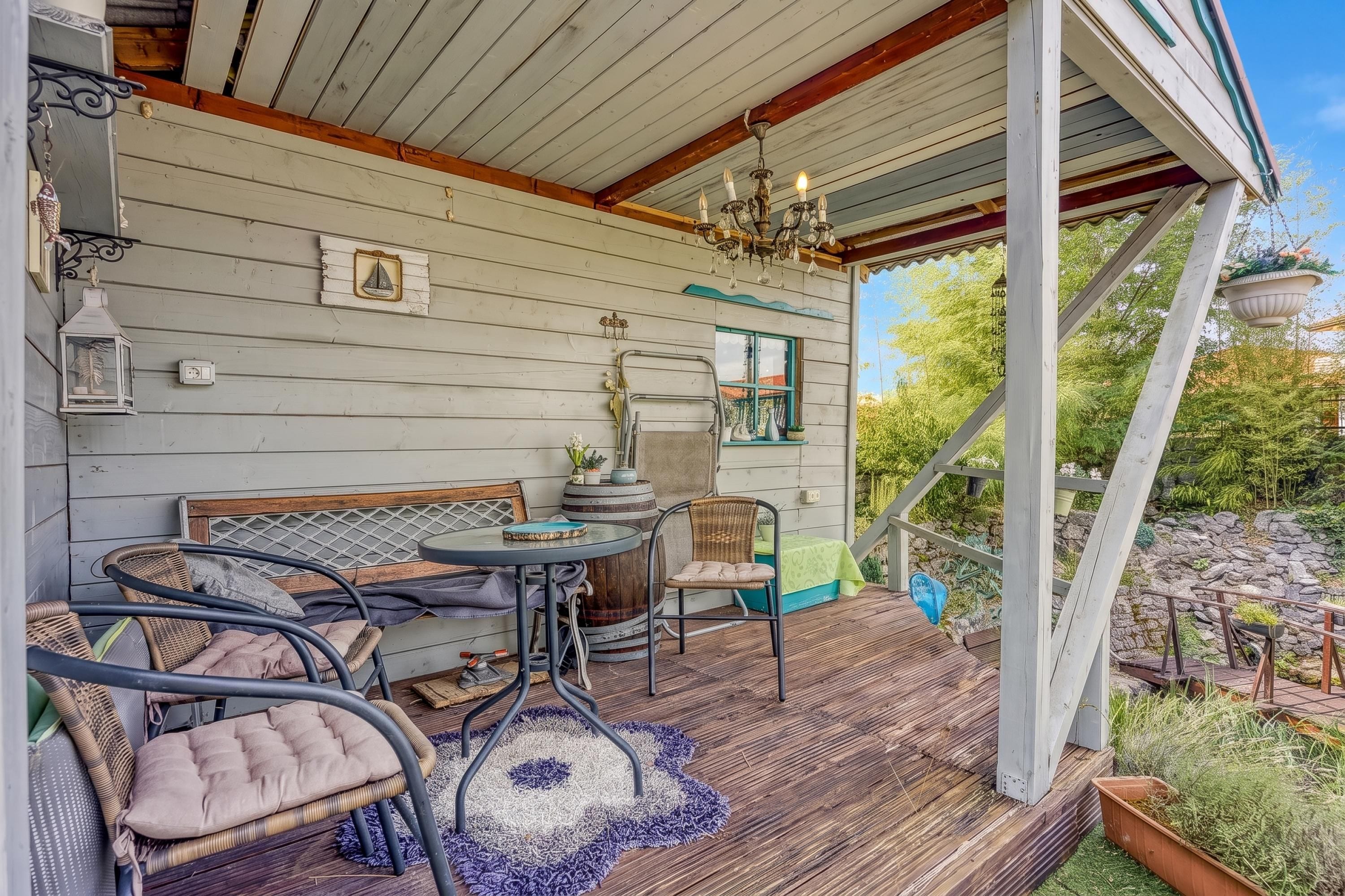 A covered outdoor seating area with a wooden deck, metal chairs, a round table, and a chandelier.