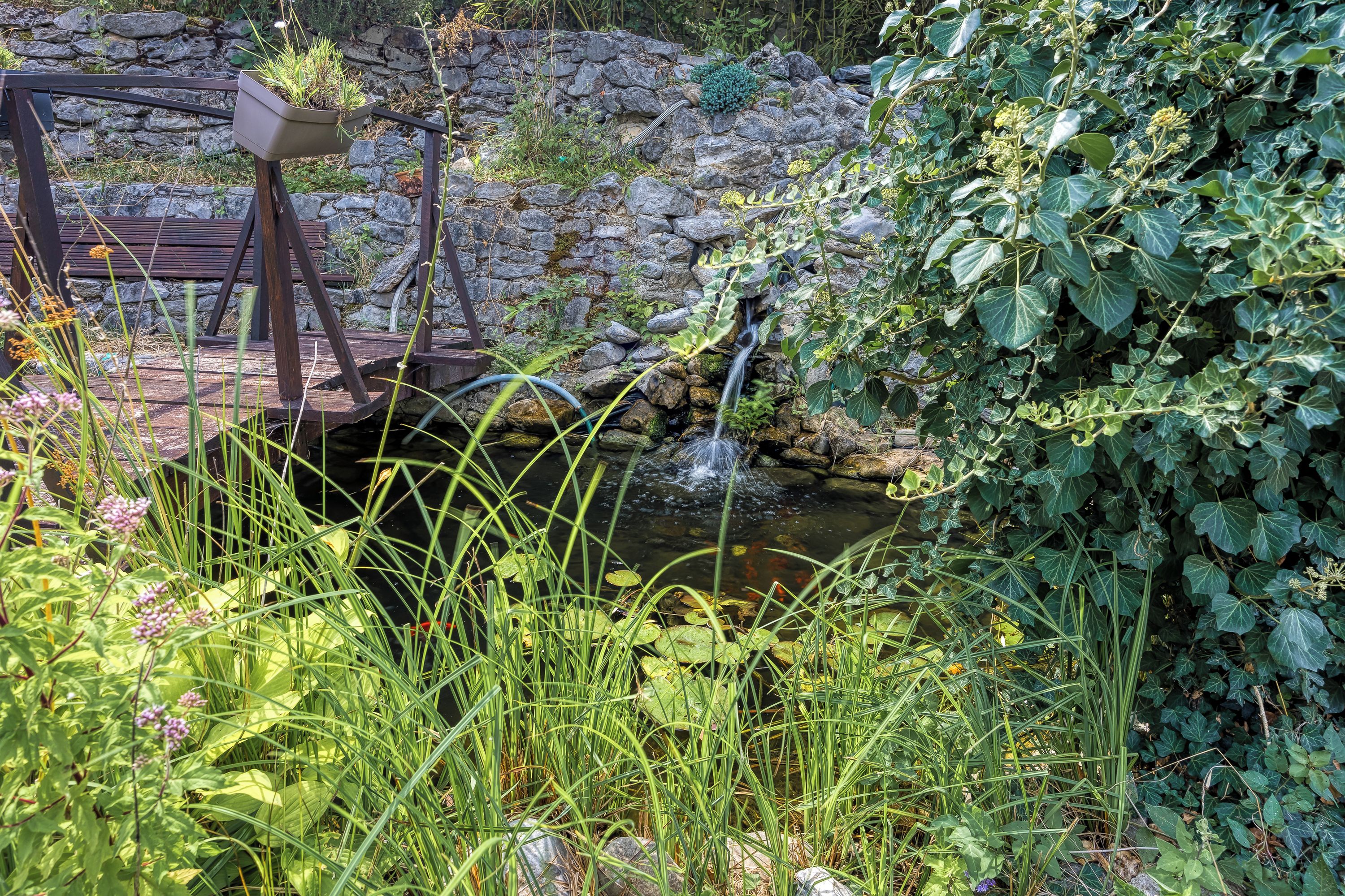 Un giardino con un muro in pietra, una panchina di legno e un piccolo ruscello.