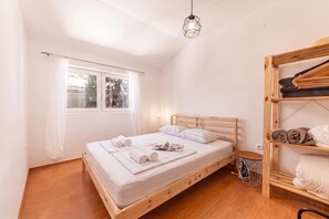 Ένα υπνοδωμάτιο με ένα ξύλινο κρεβάτι, ένα παράθυρο με κουρτίνες, ένα φωτιστικό που κρέμεται από την οροφή και ένα ξύλινο ράφι με πετσέτες.
