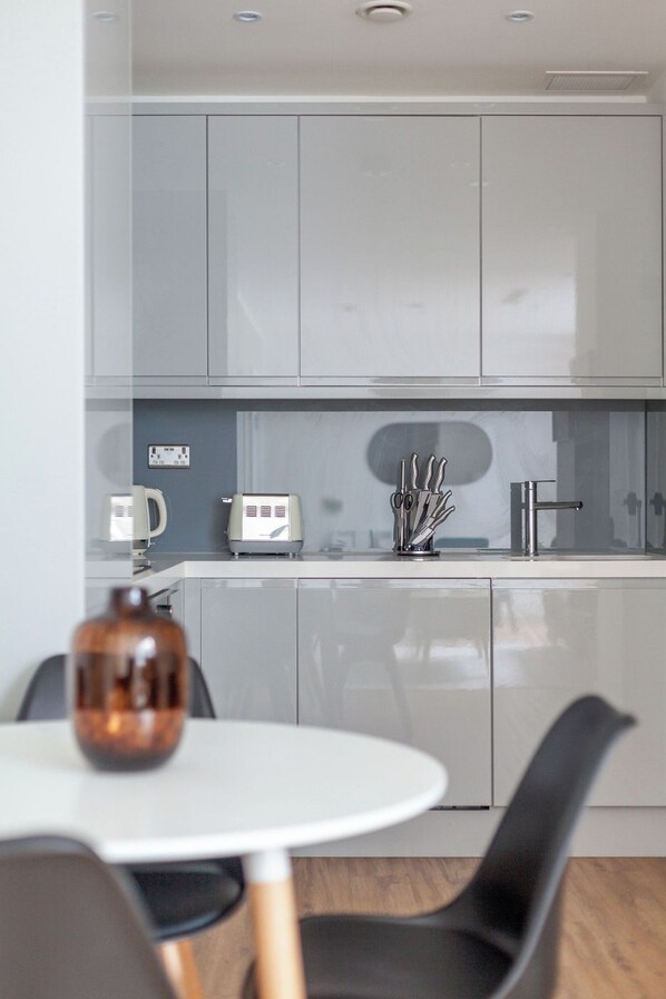 A modern kitchen with grey cabinets, a white countertop, and a coffee maker. There is a dining table with chairs in the foreground. - The Ealing Luxury Flat (London)