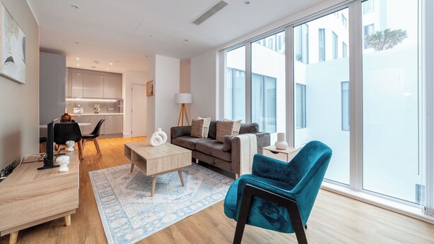 A modern living room with a sofa, coffee table, and a teal armchair. There is a dining area with a table and chairs, and a kitchen in the background. - The Ealing Luxury Flat (London)