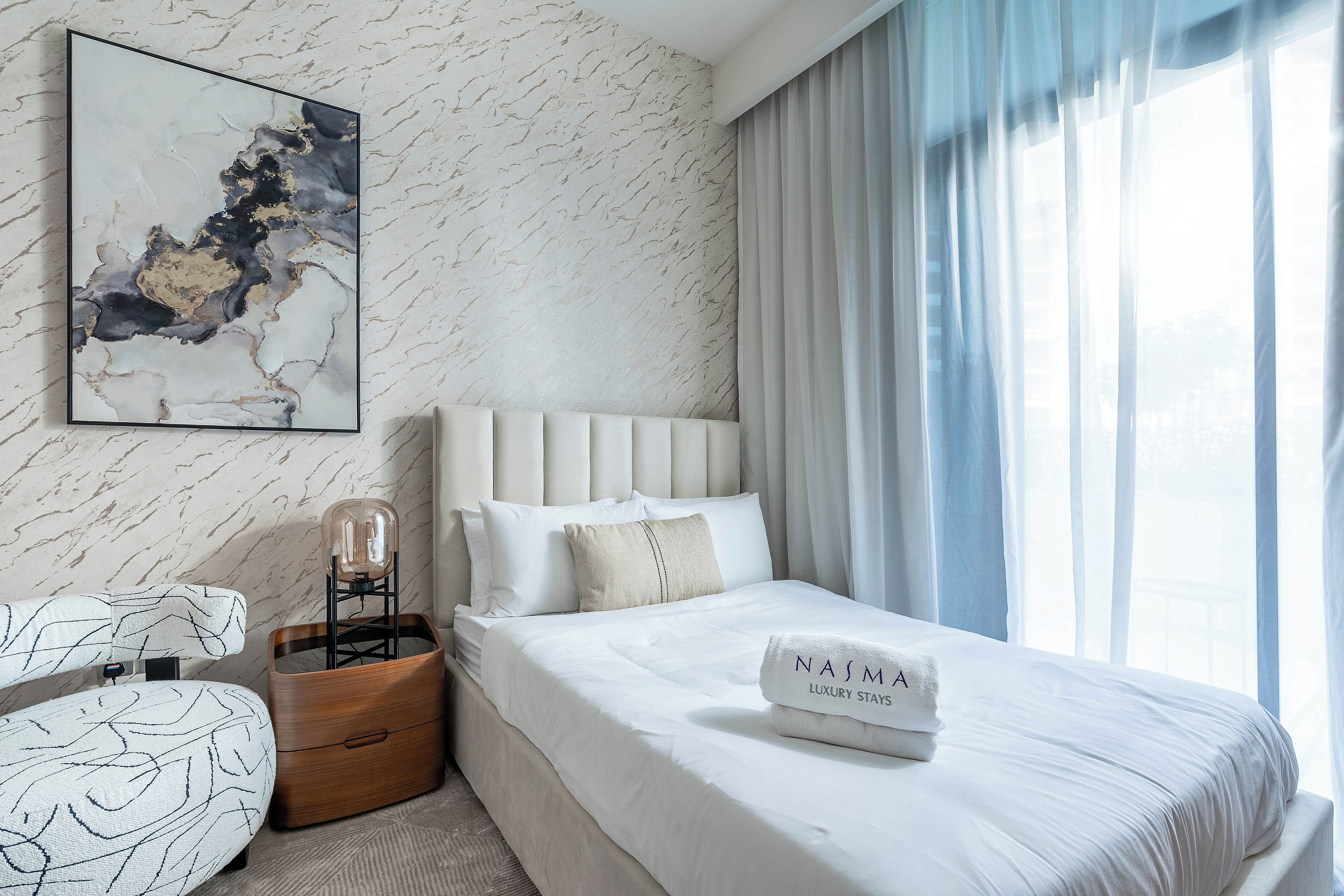 A modern hotel room with a marble-patterned wall, a large bed with white bedding, a bedside table with a lamp, and a framed abstract artwork.