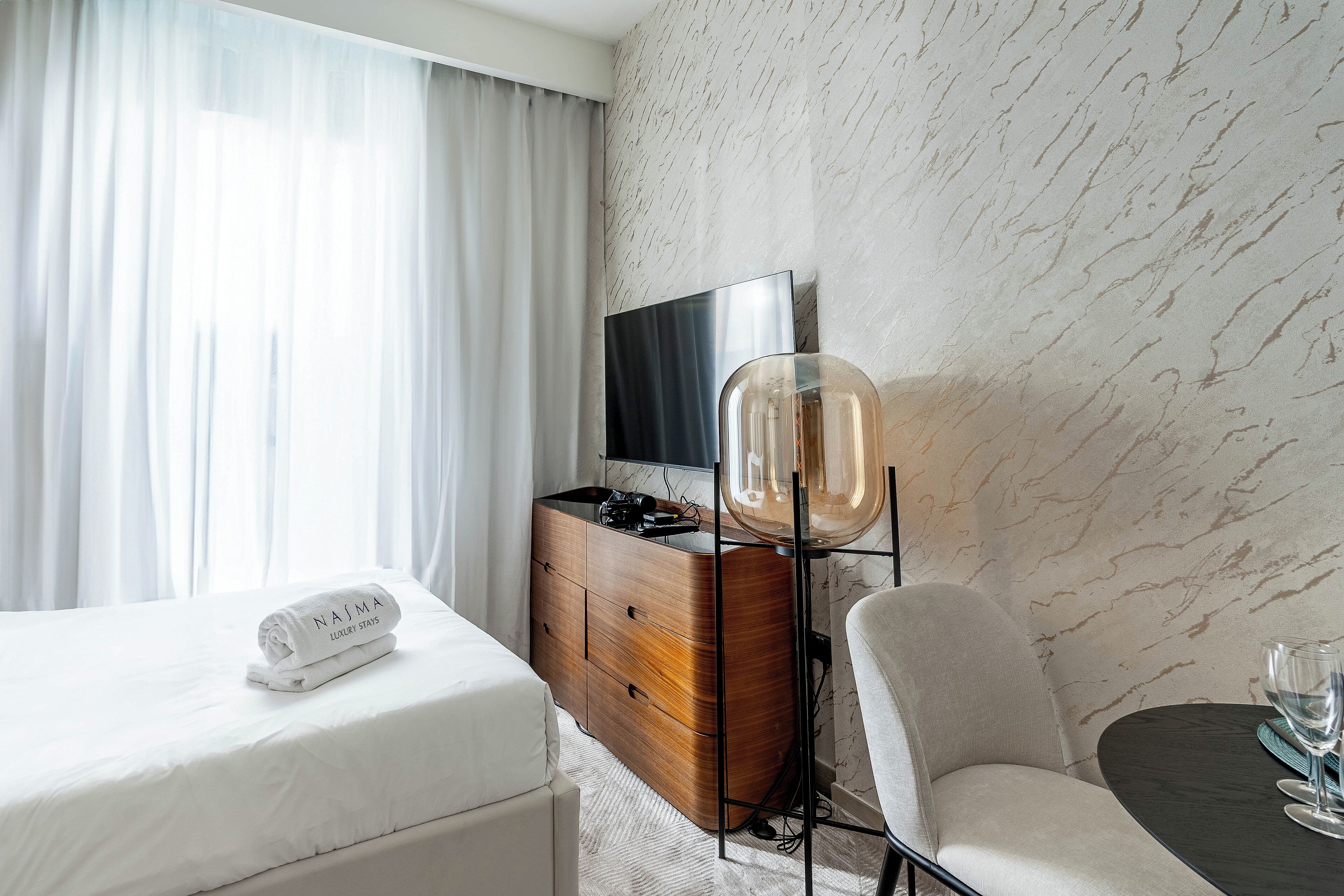 A hotel room with a bed, a wooden dresser, a TV, a modern lamp, a chair, and a small table with glasses.