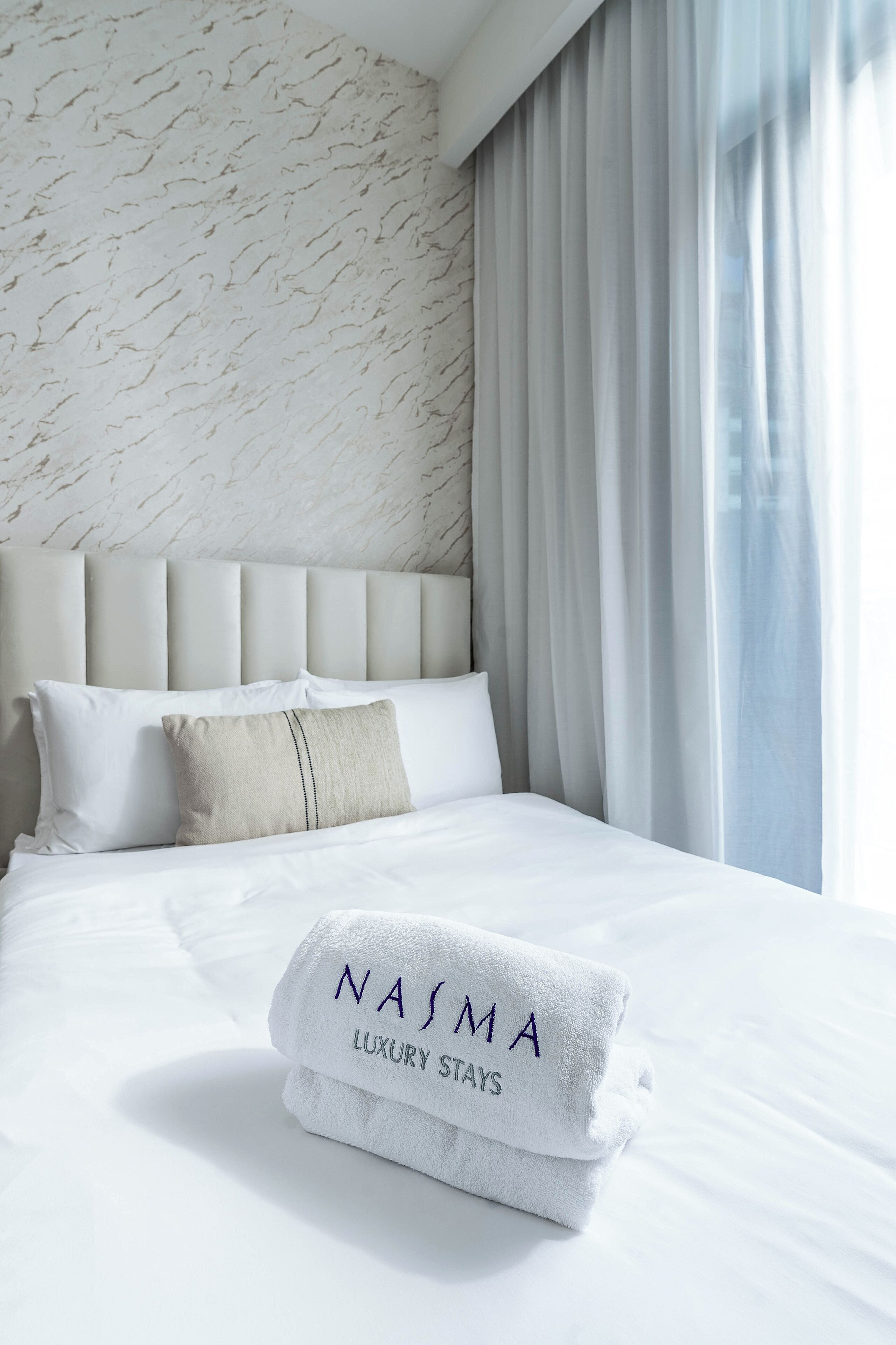 Ein ordentlich bezogenes Bett mit einem gefalteten Handtuch mit der Aufschrift "NASIMA LUXURY STAYS" und einem Kopfstück im Marmor-Design.