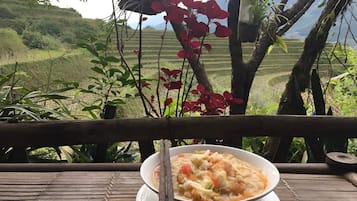 Un bol de nourriture sur une table en bois, avec vue sur des champs en terrasses et des arbres.