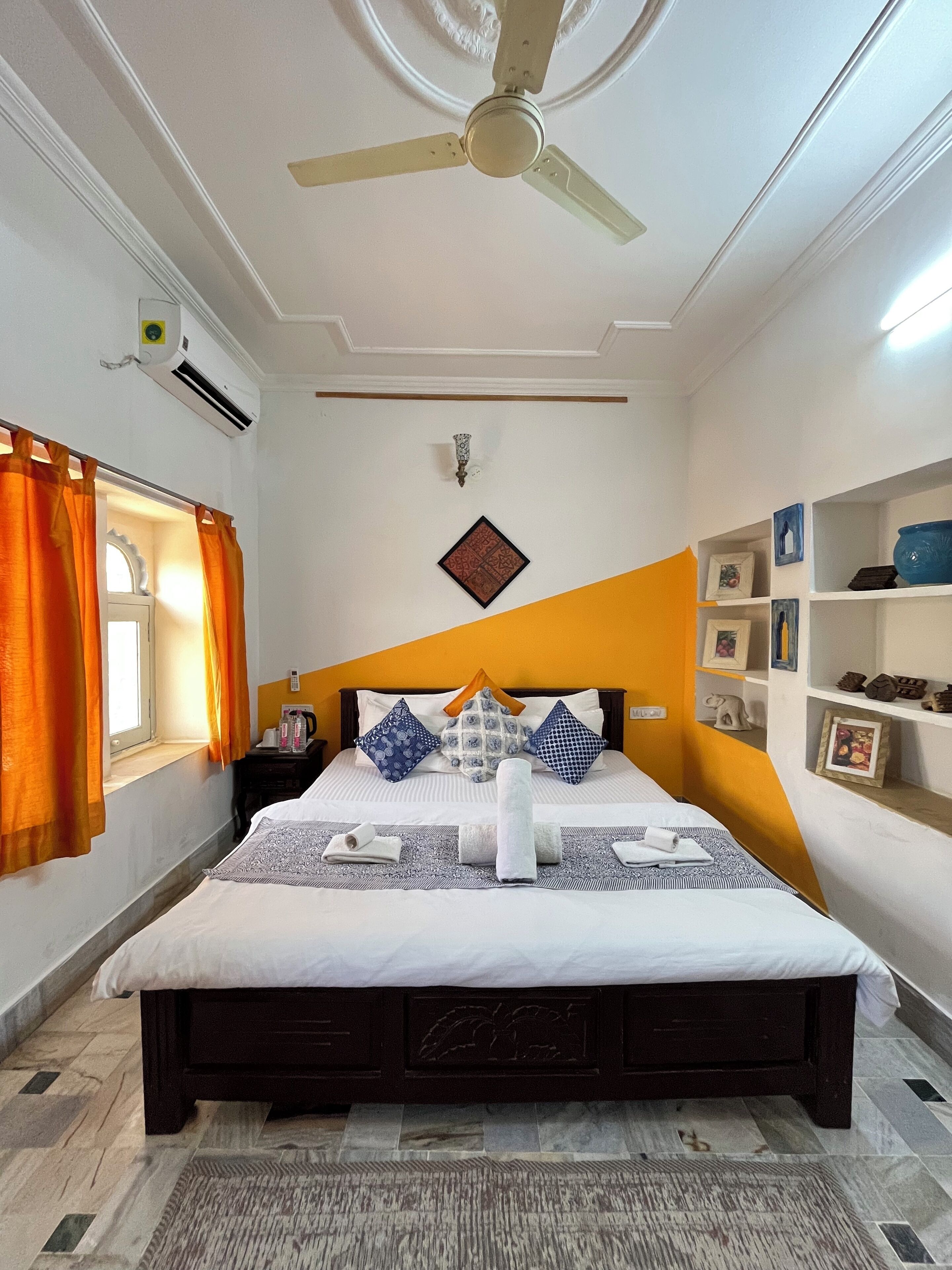 A neatly arranged hotel room with a bed, a ceiling fan, a wall-mounted air conditioner, and a window with orange curtains.