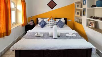A neatly arranged hotel room with a bed, a ceiling fan, a wall-mounted air conditioner, and a window with orange curtains.
