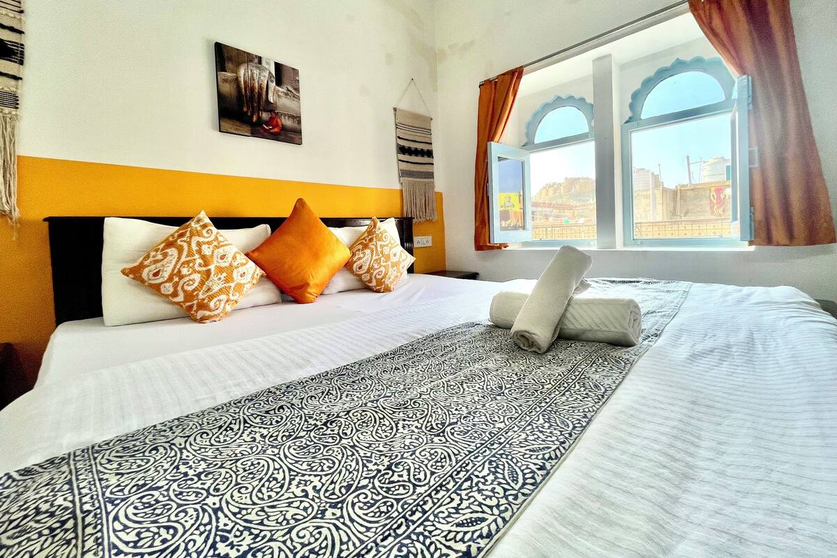 A hotel room with a bed, patterned bedspread, pillows, a painting on the wall, and a window with curtains.