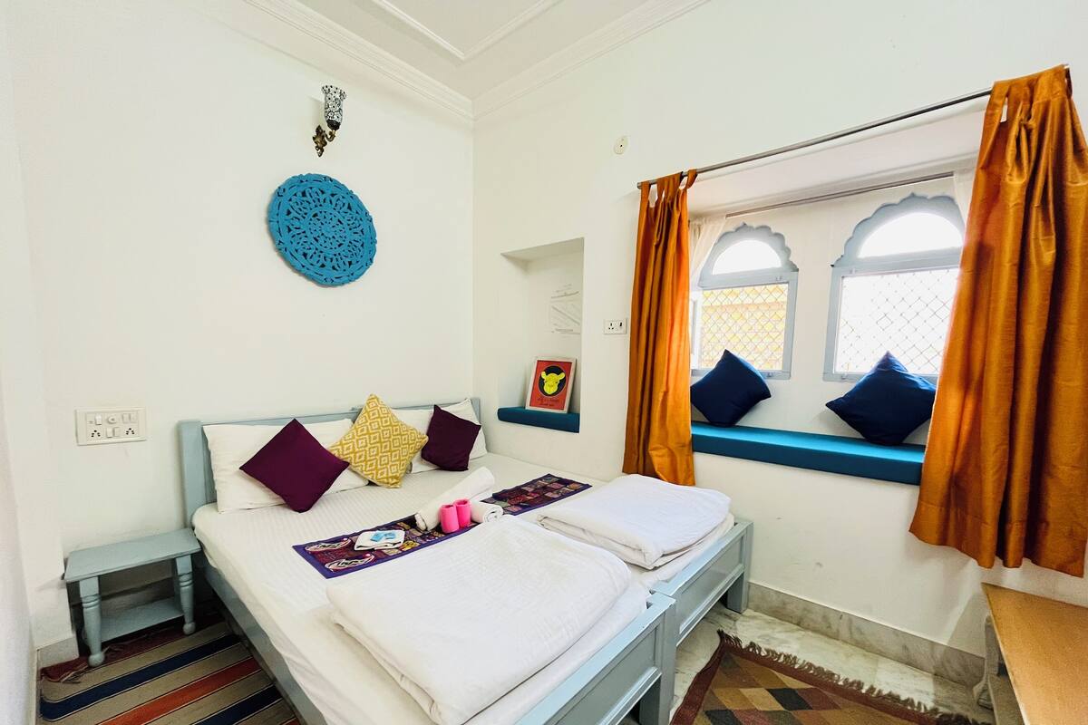 A neatly made double bed with white linens, a colorful striped rug, and a window with orange curtains.