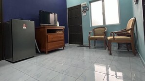Una habitación con ventilador de techo, televisión sobre un mueble de madera, refrigerador, dos sillas de madera y una ventana con persianas.
