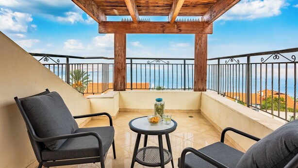 Een balkon met een houten pergola, twee stoelen, een klein rond tafeltje en uitzicht op zee.