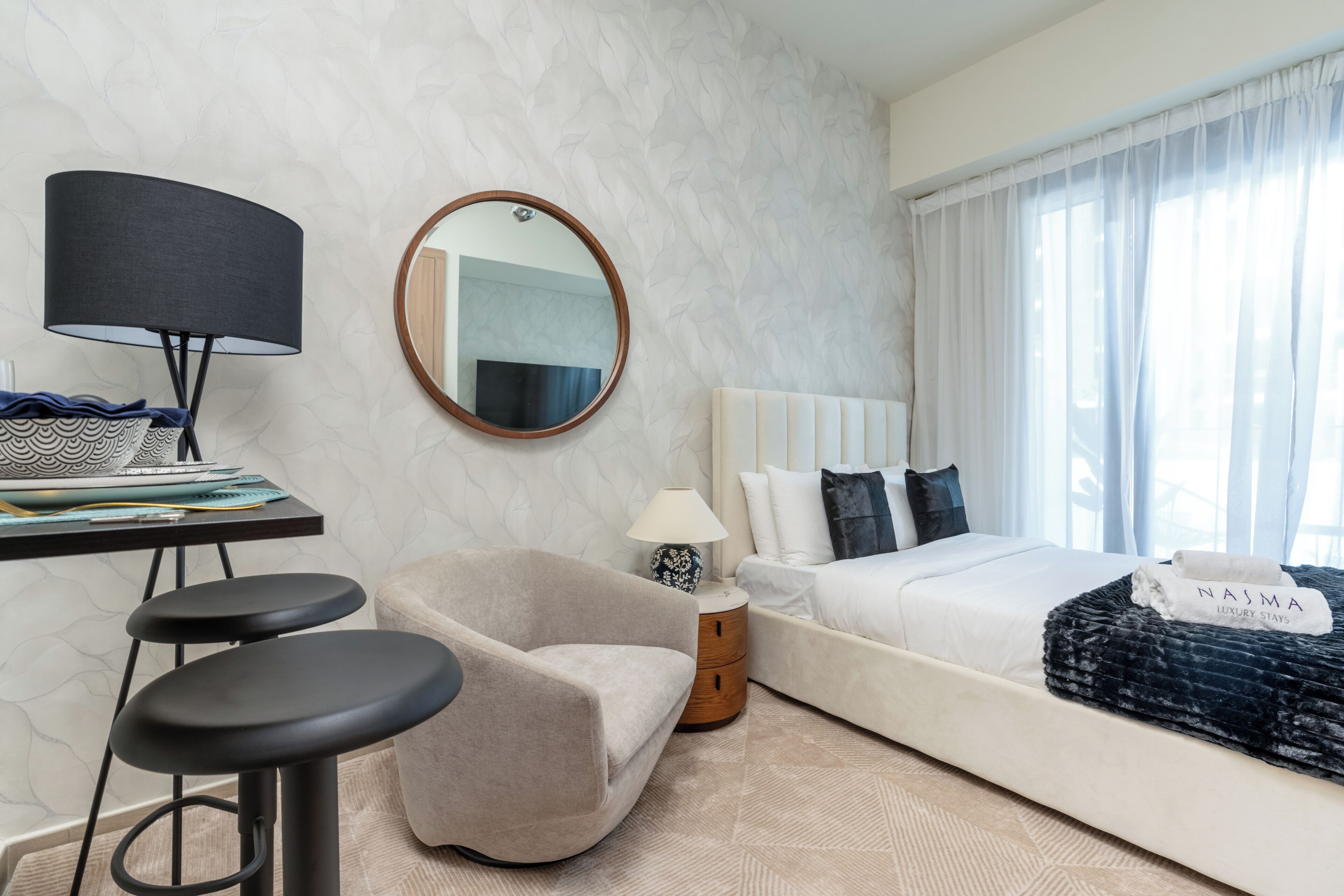 Et moderne hotelværelse med seng, skrivebord med lampe, stol og et rundt spejl.