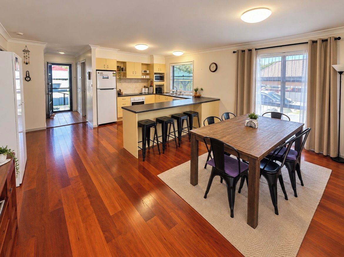 A modern kitchen with a central island, a dining area with a wooden table and chairs, and a view of the outside through large windows.