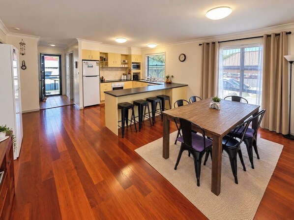 A modern kitchen with a central island, a dining area with a wooden table and chairs, and a view of the outside through large windows. - Boat Access with short Walk to River, Pub and Bowling club. (Harrington)