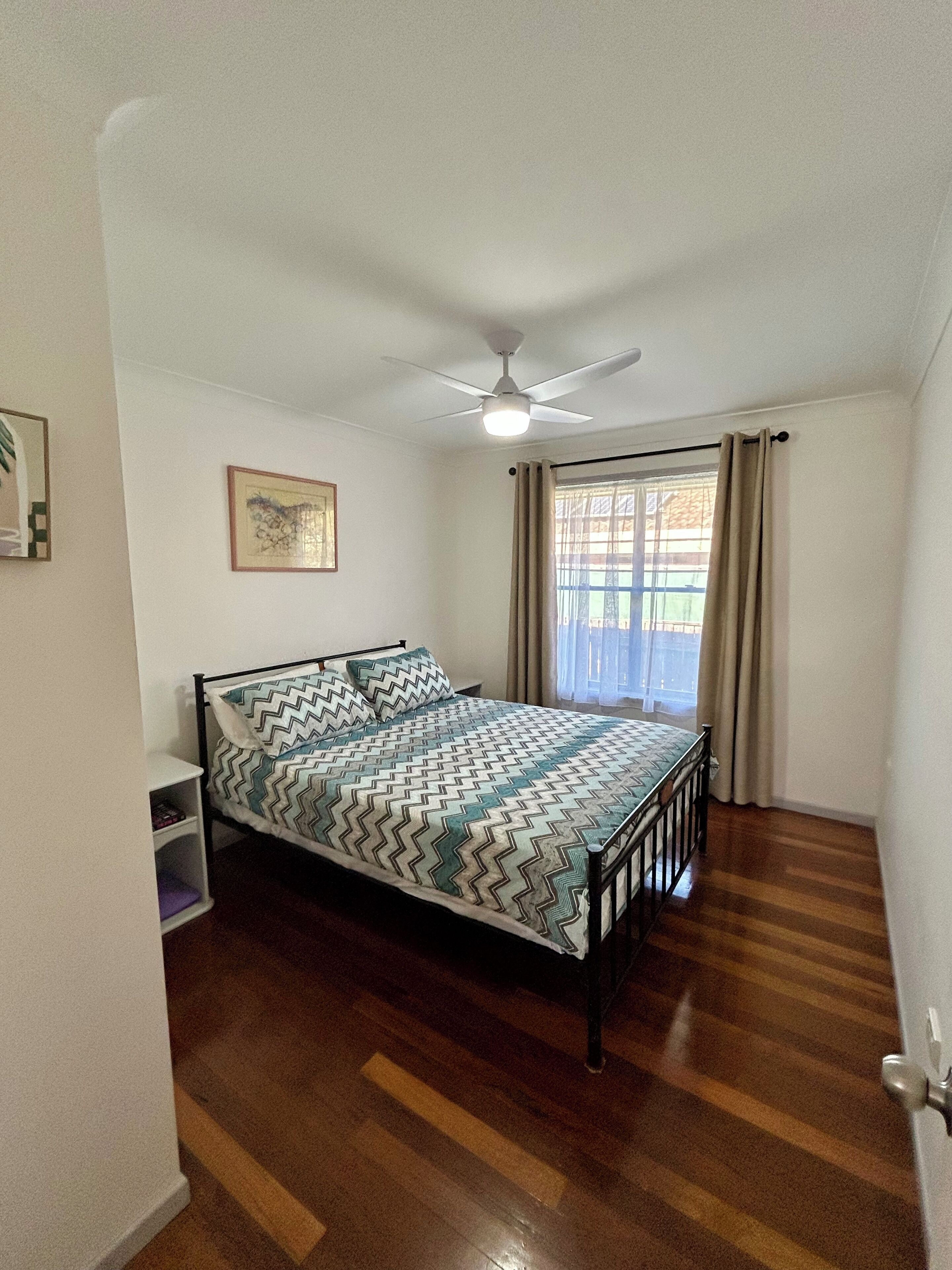 A bedroom with a bed, a ceiling fan, a window with curtains, and a wooden floor.
