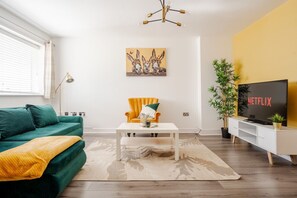 Una sala de estar moderna con un sofá verde, un sillón amarillo, una mesa de centro blanca, un cuadro de burros, un televisor que muestra 'NETFLIX' y una planta de bambú.