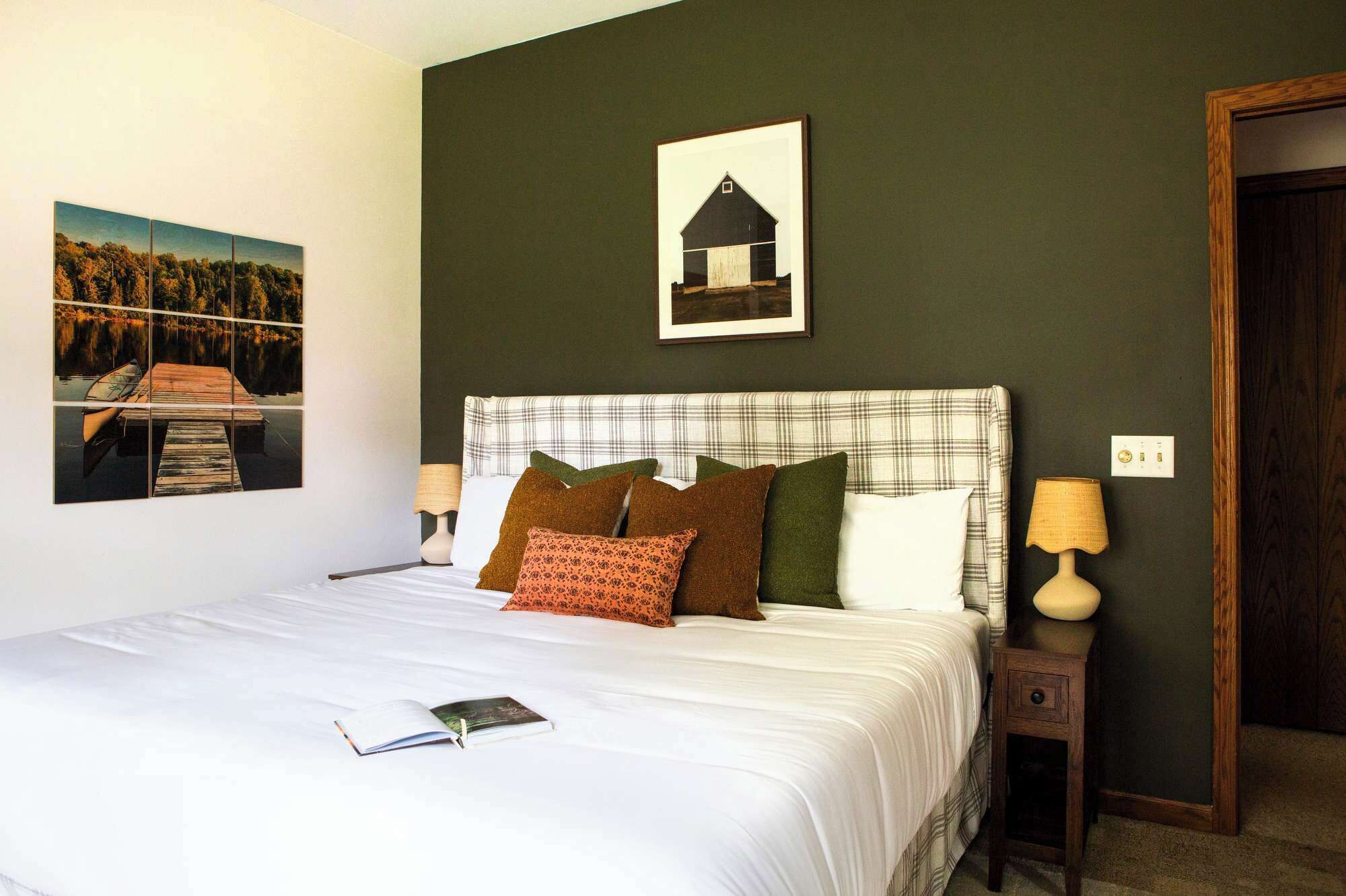 A neatly made bed with a white comforter and multiple pillows, a bedside table with a lamp, and a framed picture of a barn on the wall.