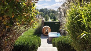 Un sentier mène à un sauna en forme de tonneau en bois et à un bain à remous situés dans un jardin aménagé.