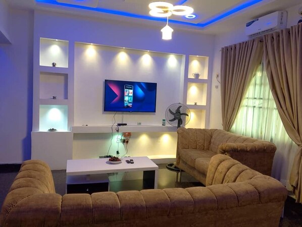 A modern living room with a flat-screen TV mounted on the wall, a sofa, a coffee table, and a ceiling fan. - 2 Bed - Sleeps 4 With Free Parking (Benin City)